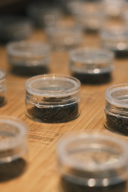 Small transparent containers are arranged on a wooden surface. Each container holds a dark substance, potentially dried leaves or herbs. The focus is on the containers in the foreground, with a gradual blur effect on those further away.