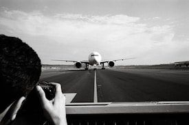 A large airplane is taxiing on a runway, central to the image, moving towards the camera. In the foreground, a photographer is capturing the scene with a camera, framing the shot through a viewfinder. The surroundings are expansive, with another airplane visible in the background to the right, and a vast, open sky above.