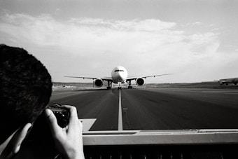 A large airplane is taxiing on a runway, central to the image, moving towards the camera. In the foreground, a photographer is capturing the scene with a camera, framing the shot through a viewfinder. The surroundings are expansive, with another airplane visible in the background to the right, and a vast, open sky above.