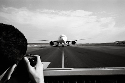 A large airplane is taxiing on a runway, central to the image, moving towards the camera. In the foreground, a photographer is capturing the scene with a camera, framing the shot through a viewfinder. The surroundings are expansive, with another airplane visible in the background to the right, and a vast, open sky above.