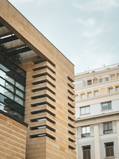 The image features a modern architectural building with a facade of tan brick and glass, juxtaposed against an older style white residential building. The sky is clear with a soft blue tone, providing a clean backdrop.