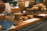 A collection of ceramic bowls and pots displayed on a wooden table in a store. The setup includes multiple pieces of pottery, each with unique designs and glazes. The background suggests a blurred setting with additional items, enhancing the warm, inviting atmosphere of the display.