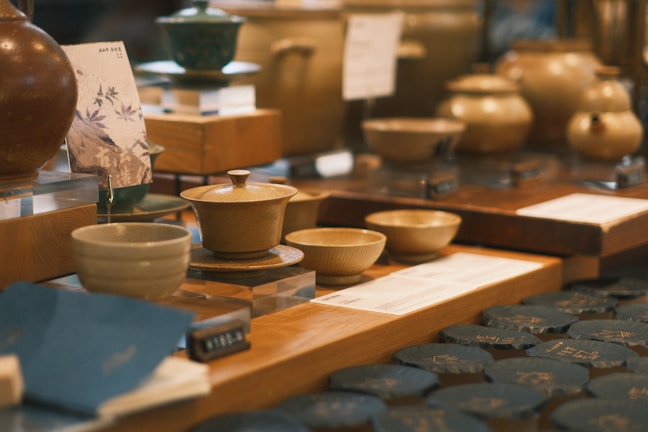 A collection of ceramic bowls and pots displayed on a wooden table in a store. The setup includes multiple pieces of pottery, each with unique designs and glazes. The background suggests a blurred setting with additional items, enhancing the warm, inviting atmosphere of the display.