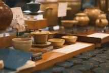 A collection of ceramic bowls and pots displayed on a wooden table in a store. The setup includes multiple pieces of pottery, each with unique designs and glazes. The background suggests a blurred setting with additional items, enhancing the warm, inviting atmosphere of the display.