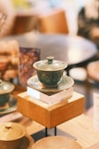 A ceramic teacup with a lid sits on a wooden pedestal in a softly lit room. The cup features a mottled green pattern, and the surroundings include blurred figures and tables, creating a tranquil atmosphere.