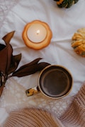A warm and cozy setup features a lit candle with a pumpkin-shaped holder, a mug filled with a hot beverage, some dried leaves, and a couple of ornamental squash. The items rest on a white, intricately patterned cloth alongside a knitted sweater, conveying a rustic, autumnal atmosphere.