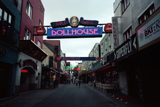 A narrow street flanked by buildings on either side, displaying colorful neon signs for clubs and bars, such as 'Dollhouse' and 'Superfly'. The street is slightly crowded with a few people walking in the distance. The mood is lively, typical of nightlife districts.