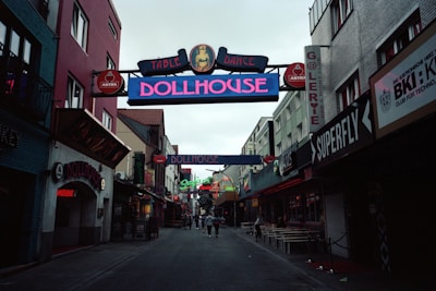 A narrow street flanked by buildings on either side, displaying colorful neon signs for clubs and bars, such as 'Dollhouse' and 'Superfly'. The street is slightly crowded with a few people walking in the distance. The mood is lively, typical of nightlife districts.