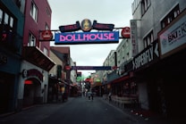 A narrow street flanked by buildings on either side, displaying colorful neon signs for clubs and bars, such as 'Dollhouse' and 'Superfly'. The street is slightly crowded with a few people walking in the distance. The mood is lively, typical of nightlife districts.