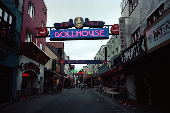 A narrow street flanked by buildings on either side, displaying colorful neon signs for clubs and bars, such as 'Dollhouse' and 'Superfly'. The street is slightly crowded with a few people walking in the distance. The mood is lively, typical of nightlife districts.