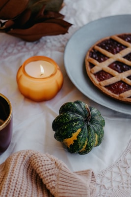 A cozy autumn setting featuring a lit candle in an orange holder, a small green pumpkin with orange patches, a knitted beige blanket, a cup with a gold rim, and a lattice-topped berry pie on a gray plate. The background includes a lace tablecloth adding to the warm and inviting atmosphere.
