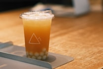 A plastic cup filled with a brownish-orange iced drink sits on a tray. The cup has a transparent lid and is partially filled with small tapioca pearls at the bottom. The drink is set on a wooden surface, and there is a geometric mountain logo printed on the cup.