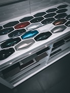 A display of various car paint color samples arranged in neat rows on a white surface. Each sample is shaped like a car body panel and showcases a different shade, ranging from dark greens and blues to reds, silvers, and blacks.
