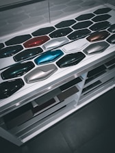 A display of various car paint color samples arranged in neat rows on a white surface. Each sample is shaped like a car body panel and showcases a different shade, ranging from dark greens and blues to reds, silvers, and blacks.