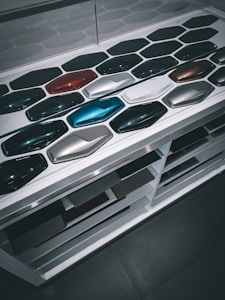 A display of various car paint color samples arranged in neat rows on a white surface. Each sample is shaped like a car body panel and showcases a different shade, ranging from dark greens and blues to reds, silvers, and blacks.
