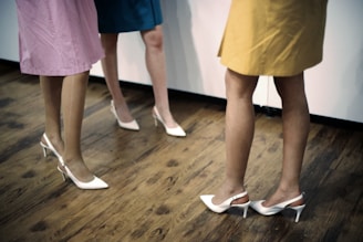 Three individuals are standing on a wooden floor, wearing colorful knee-length skirts in pink, blue, and yellow, along with white high-heeled shoes. The focus is on their legs and shoes.