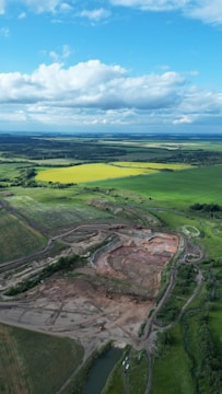 An aerial view of a vast landscape with a mixture of cultivated fields and undeveloped land. The foreground showcases a large excavation site with visible construction vehicles and dirt tracks. Surrounding the site are lush green fields, with some patches of vibrant yellow. In the distance, the horizon is filled with more fields and a line of dense trees. The sky overhead is mostly clear with some large, fluffy clouds.