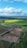 An aerial view of a vast landscape with a mixture of cultivated fields and undeveloped land. The foreground showcases a large excavation site with visible construction vehicles and dirt tracks. Surrounding the site are lush green fields, with some patches of vibrant yellow. In the distance, the horizon is filled with more fields and a line of dense trees. The sky overhead is mostly clear with some large, fluffy clouds.
