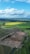 An aerial view of a vast landscape with a mixture of cultivated fields and undeveloped land. The foreground showcases a large excavation site with visible construction vehicles and dirt tracks. Surrounding the site are lush green fields, with some patches of vibrant yellow. In the distance, the horizon is filled with more fields and a line of dense trees. The sky overhead is mostly clear with some large, fluffy clouds.