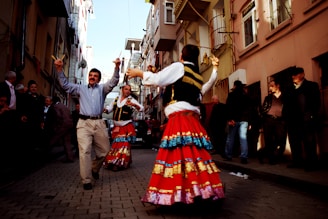 A lively street scene with people dancing in traditional attire. The foreground features two dancers wearing colorful costumes with bright red skirts adorned with blue and gold embellishments. Around them, other individuals are gathered, some watching the performance and others walking by. The setting is a narrow, urban alley lined with tall buildings, creating a festive and vibrant atmosphere.