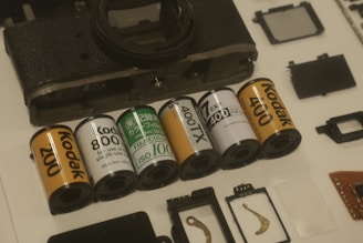 A disassembled camera surrounded by several rolls of film, including Kodak and Fujicolor brands, arranged neatly on a surface. Additional small components and parts are scattered around, such as metal pieces and circuit boards.