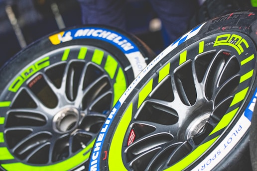 Two racing tires with vibrant yellow-green and blue accents are shown, featuring detailed tread patterns and the brand name Michelin visible on the sidewall. The rims are sleek and metallic with a modern design.