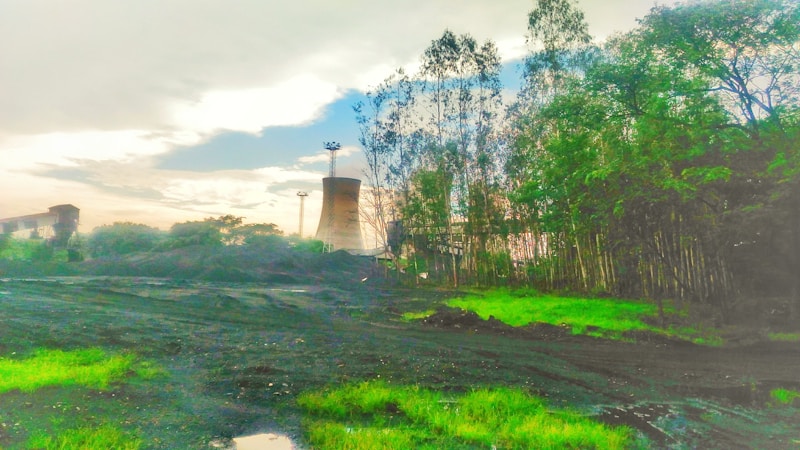 A landscape featuring a cooling tower in the background surrounded by greenery and trees. The foreground consists of dark soil and patches of bright green grass, indicating a mix of industrial and natural elements. The sky is overcast with clouds, creating a contrasting backdrop to the scene.