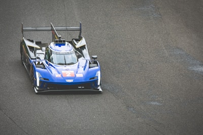 A blue racing car with a sleek aerodynamic design is on a race track. The car displays the number 2 in bold red on its front. It features prominent spoilers and branding on its body, including the name Cadillac. The texture of the asphalt is visible, suggesting speed and precision typical in a race environment.
