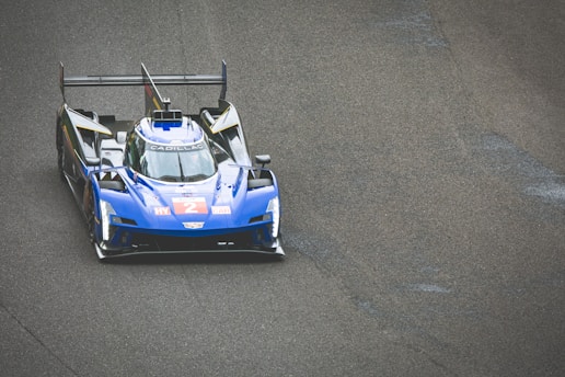 A blue racing car with a sleek aerodynamic design is on a race track. The car displays the number 2 in bold red on its front. It features prominent spoilers and branding on its body, including the name Cadillac. The texture of the asphalt is visible, suggesting speed and precision typical in a race environment.