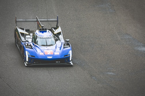 A blue racing car with a sleek aerodynamic design is on a race track. The car displays the number 2 in bold red on its front. It features prominent spoilers and branding on its body, including the name Cadillac. The texture of the asphalt is visible, suggesting speed and precision typical in a race environment.