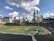 A baseball field with a green, well-maintained outfield and clay infield surrounded by empty stadium seats. A few players and staff are on the field preparing, with batting cages set up near home plate. The backdrop includes a city skyline with tall buildings under a partly cloudy sky.