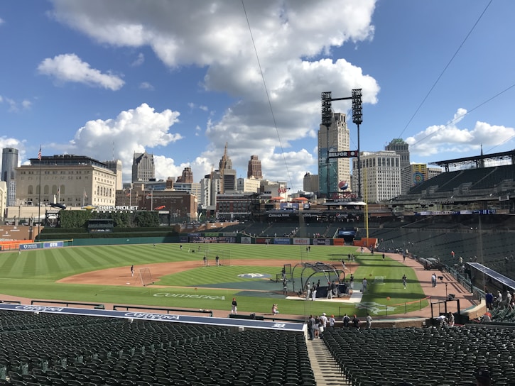 A baseball field with a green, well-maintained outfield and clay infield surrounded by empty stadium seats. A few players and staff are on the field preparing, with batting cages set up near home plate. The backdrop includes a city skyline with tall buildings under a partly cloudy sky.