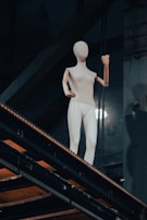 A mannequin with a wooden head and arms positioned in a casual stance on a metal stairway in an industrial setting. The lighting highlights the clean and simplistic design of the mannequin against the dark background.