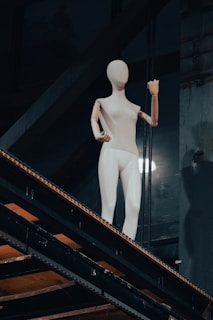 A mannequin with a wooden head and arms positioned in a casual stance on a metal stairway in an industrial setting. The lighting highlights the clean and simplistic design of the mannequin against the dark background.