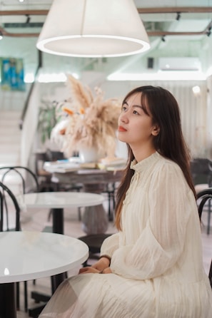 A woman in a flowing white blouse and tailored trousers standing in a bright, airy cafe with plants.