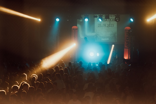 A vibrant concert scene with a large crowd of people gathered in front of a lit stage. The atmosphere is lively with colorful lights, including blue and yellow beams shining over the audience. On stage, there are DJ equipment and visual displays showing a date and event details.