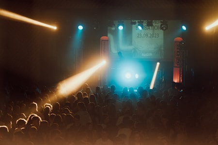 A vibrant concert scene with a large crowd of people gathered in front of a lit stage. The atmosphere is lively with colorful lights, including blue and yellow beams shining over the audience. On stage, there are DJ equipment and visual displays showing a date and event details.