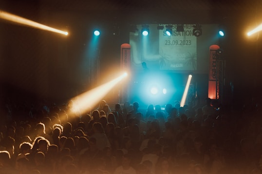 A vibrant concert scene with a large crowd of people gathered in front of a lit stage. The atmosphere is lively with colorful lights, including blue and yellow beams shining over the audience. On stage, there are DJ equipment and visual displays showing a date and event details.
