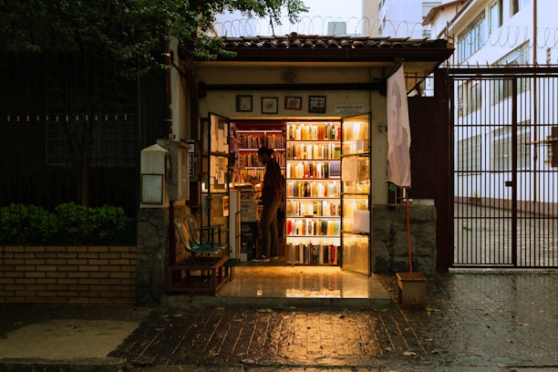 A small, cozy bookstore at night with warm lighting illuminating the books on the shelves. The store is situated in a narrow entrance with a person standing inside, browsing or arranging the books. Outside, the environment is slightly dim, creating a contrast with the glow from the bookstore. The wet pavement suggests a recent rain.
