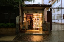 A small, cozy bookstore at night with warm lighting illuminating the books on the shelves. The store is situated in a narrow entrance with a person standing inside, browsing or arranging the books. Outside, the environment is slightly dim, creating a contrast with the glow from the bookstore. The wet pavement suggests a recent rain.