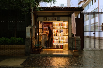 A small, cozy bookstore at night with warm lighting illuminating the books on the shelves. The store is situated in a narrow entrance with a person standing inside, browsing or arranging the books. Outside, the environment is slightly dim, creating a contrast with the glow from the bookstore. The wet pavement suggests a recent rain.