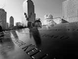 The image displays a close-up of a memorial with engraved names, set against the backdrop of a cityscape with several tall buildings under a clear sky. The perspective highlights the reflective surface of the memorial.