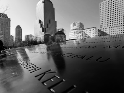 The image displays a close-up of a memorial with engraved names, set against the backdrop of a cityscape with several tall buildings under a clear sky. The perspective highlights the reflective surface of the memorial.