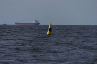 A large cargo ship sails on a vast body of water with a bright yellow navigation buoy floating in the foreground. The water appears dark and choppy under a cloudy sky.