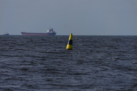 A large cargo ship sails on a vast body of water with a bright yellow navigation buoy floating in the foreground. The water appears dark and choppy under a cloudy sky.