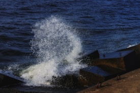 Waves crash energetically against a series of concrete barriers at the edge of a body of water. The splash creates a striking spray of water droplets against the deep blue of the ocean.