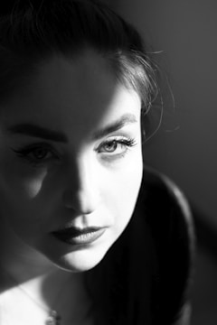 A black and white portrait of a person with dramatic lighting creating a shadow across the face. The light highlights the eyes, eyebrows, and lips, adding contrast and intensity to the expression.