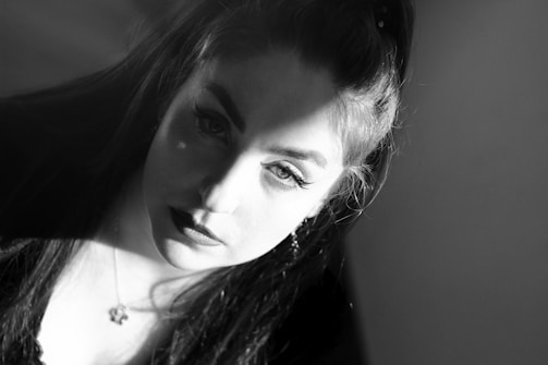A black and white portrait of a woman looking directly at the camera with a serious expression. Soft lighting creates shadows across her face, highlighting her features such as the dark lipstick and well-defined eyebrows. Her dark hair frames her face and she wears a pendant on a necklace.