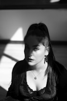 A woman with long hair tied in a high ponytail is seated and gazing directly at the viewer. Her makeup is striking, with bold eyeliner, and she wears a dark top with lace detailing. The lighting is dramatic, creating sharp contrasts and deep shadows on her face and surroundings.