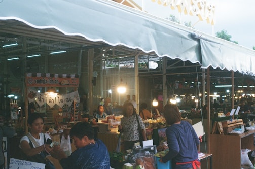 A busy market scene with several vendors and shoppers. Stalls are filled with various goods and products, and there are bright lights creating a warm, lively atmosphere. Signs with text in a Southeast Asian script hang from the stalls, adding to the market ambiance.